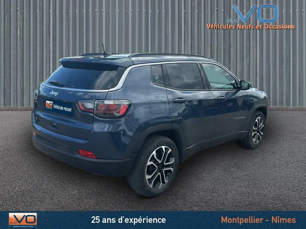 Jeep Compass bleu 3/4 arrière droit, avec barres de toit et jantes 18 pouces visibles, stationné devant un mur métallique.