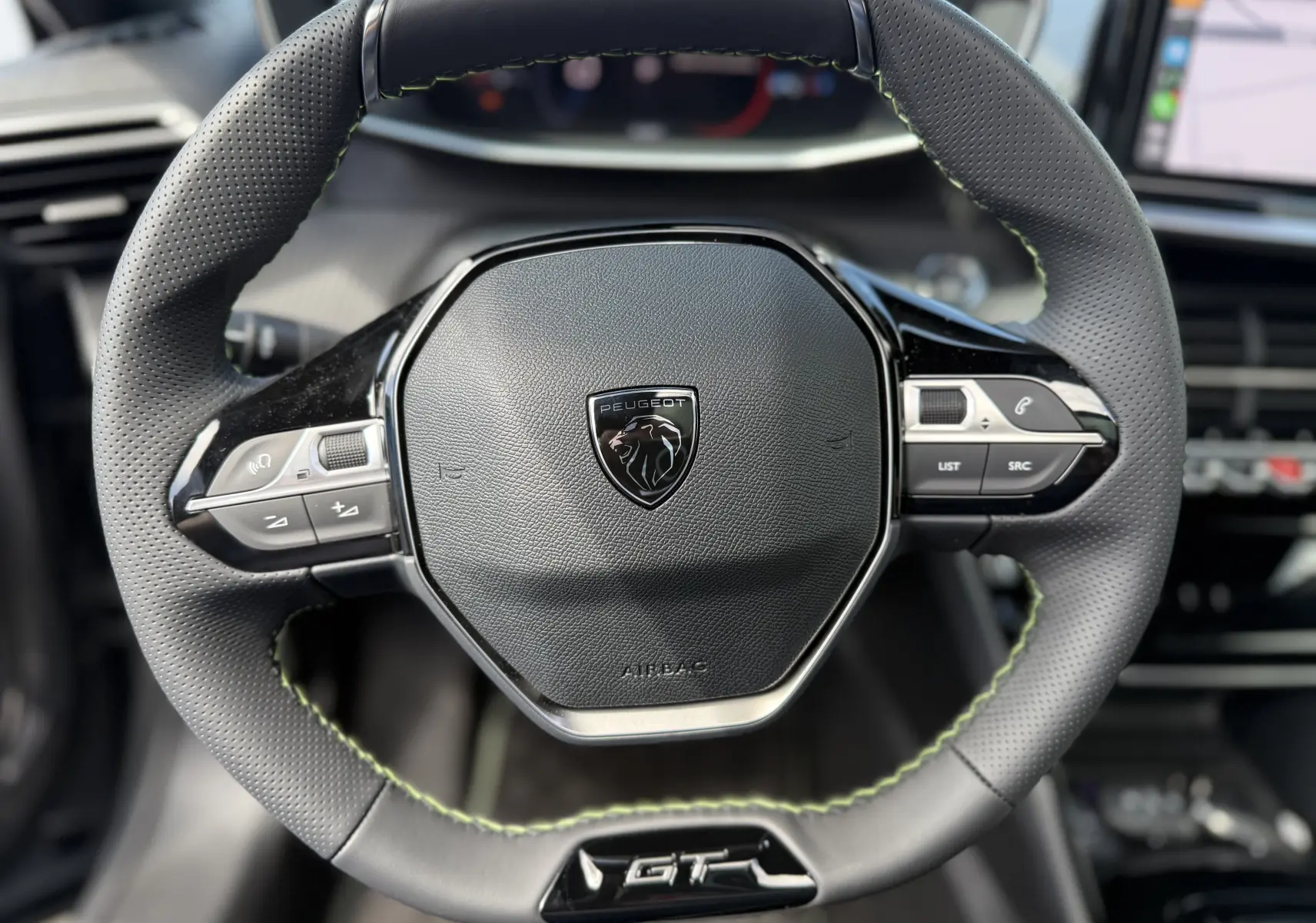Gros plan sur le volant cuir perforé noir avec surpiqûres vertes du Peugeot 2008 Hybrid 145ch GT, tableau de bord en arrière-plan flou.