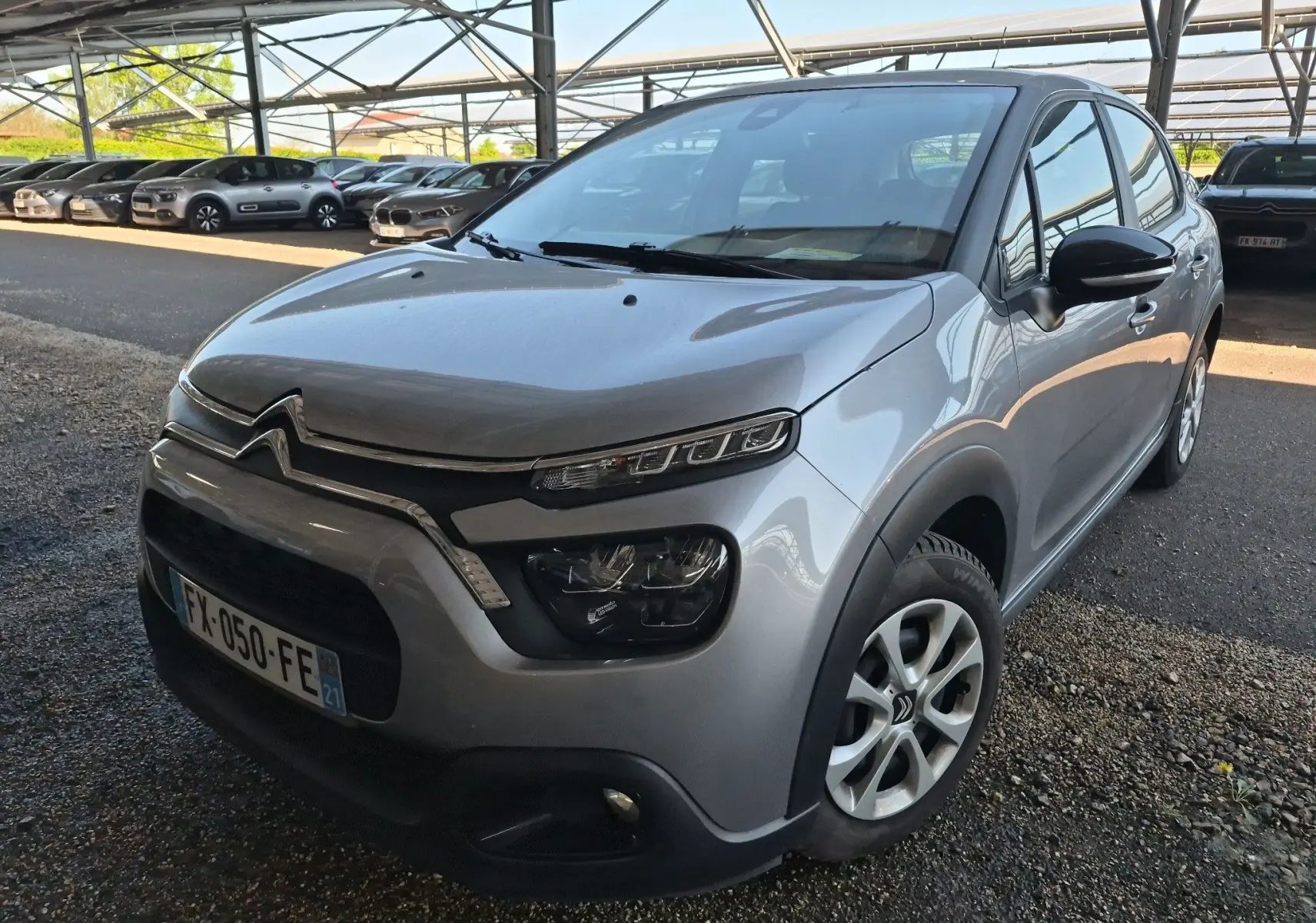 Citroën C3 gris Artense vue 3/4 avant droit, avec phares distinctifs et calandre chromée sous abri.