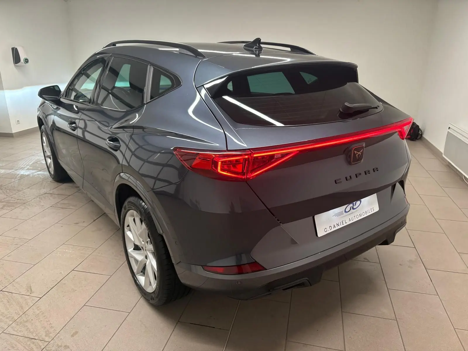 Vue 3/4 arrière droite du CUPRA Formentor gris magnétique 2024 avec feux arrière allumés et hayon fermé en intérieur.