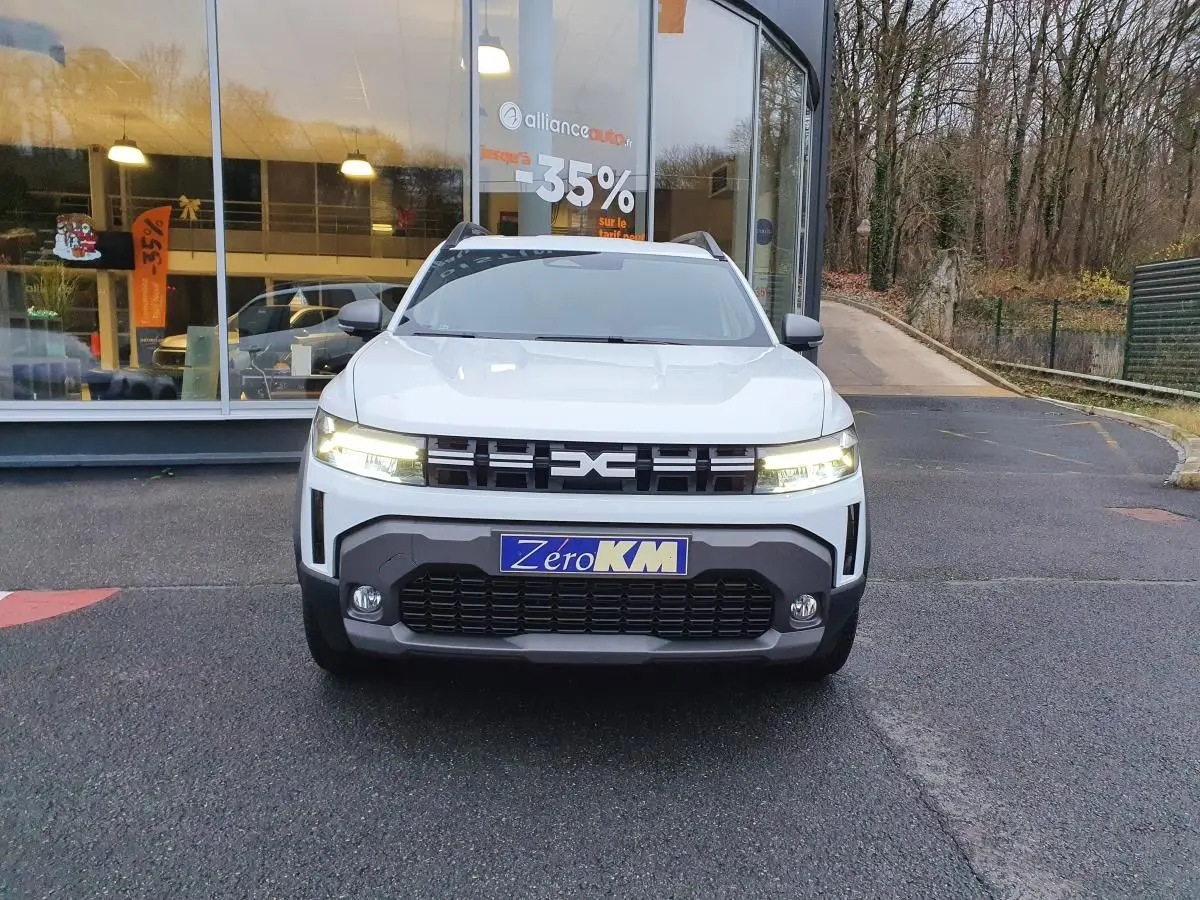 Vue frontale d’un Dacia Duster blanc glacier 2025 avec calandre noire et phares allumés devant un bâtiment vitré.