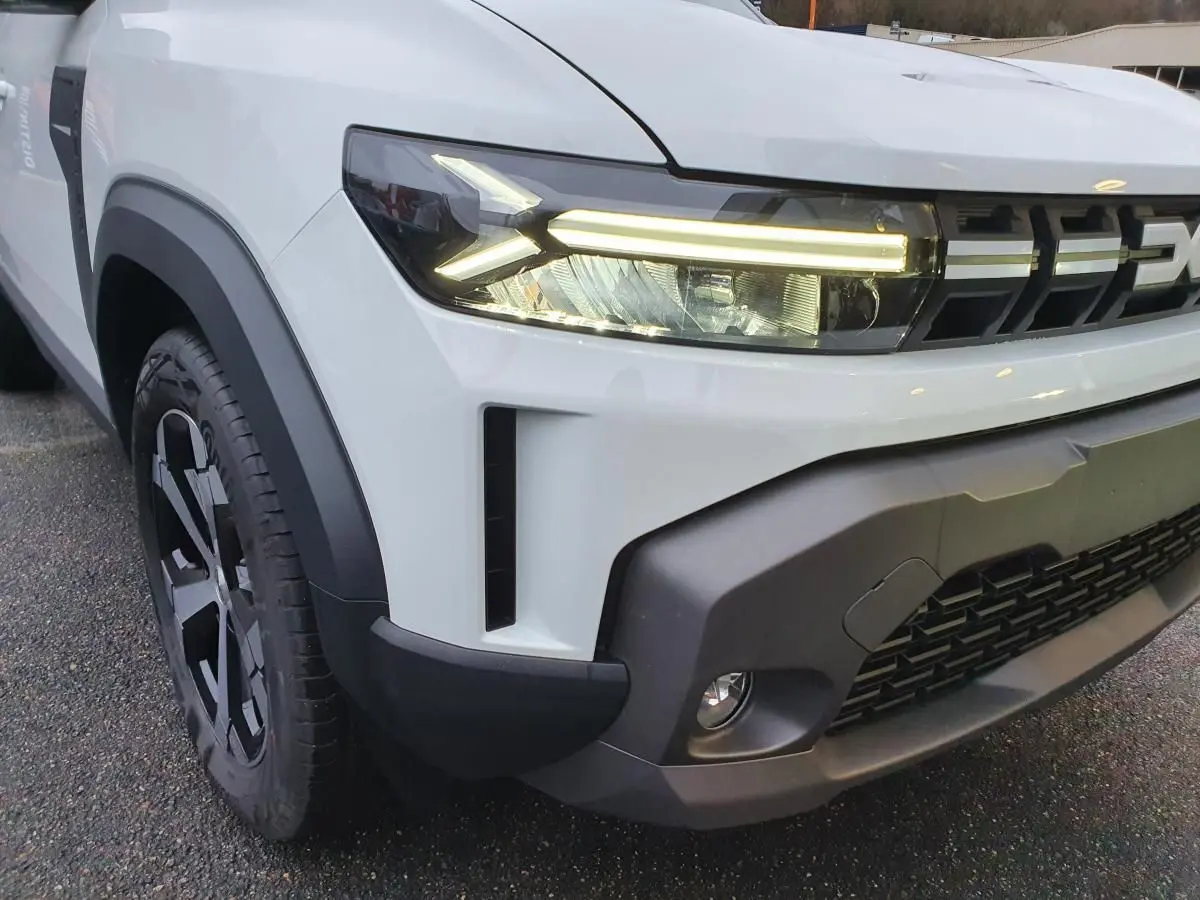 Gros plan sur l’avant gauche du Dacia Duster blanc glacier 2025, mettant en valeur les phares LED et la calandre noire.