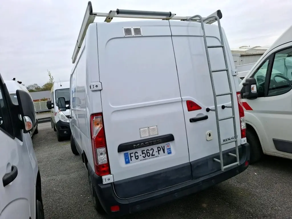 Vue arrière d’un Renault Master blanc avec galerie et échelle métallique, stationné entre deux autres véhicules utilitaires.