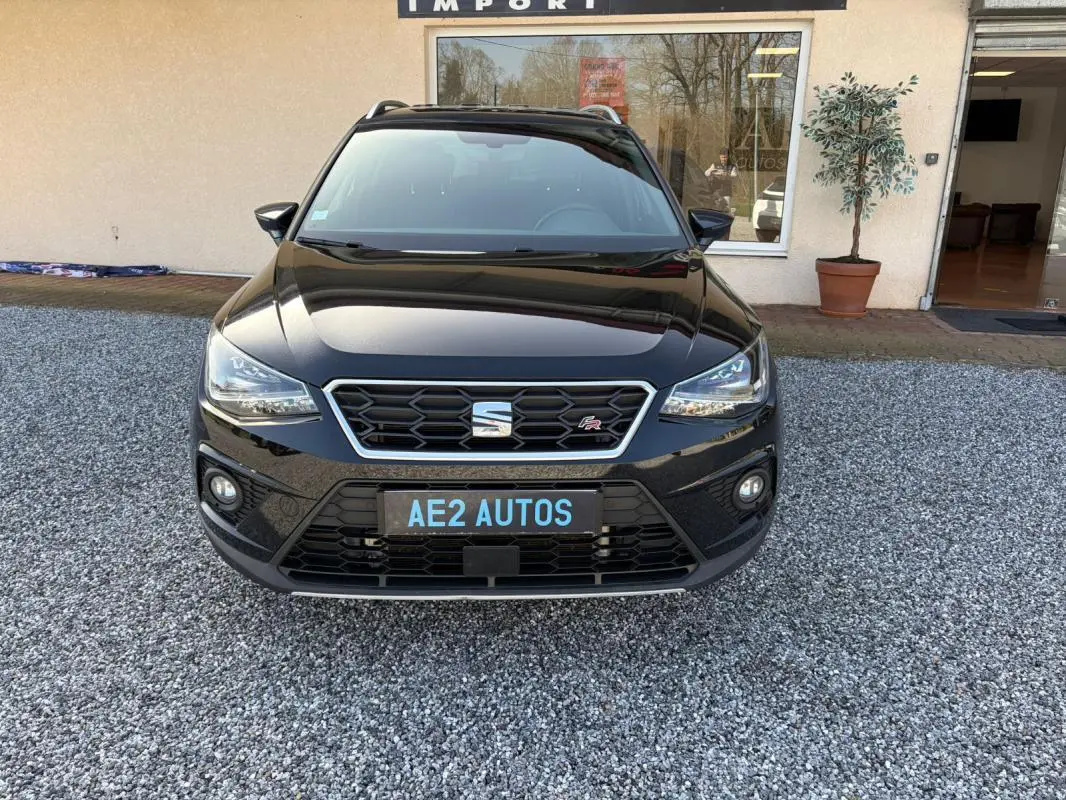 Vue frontale d'un SEAT Arona noir 2020 avec badge FR et calandre hexagonale, stationné sur un sol gravillonné devant un bâtiment.