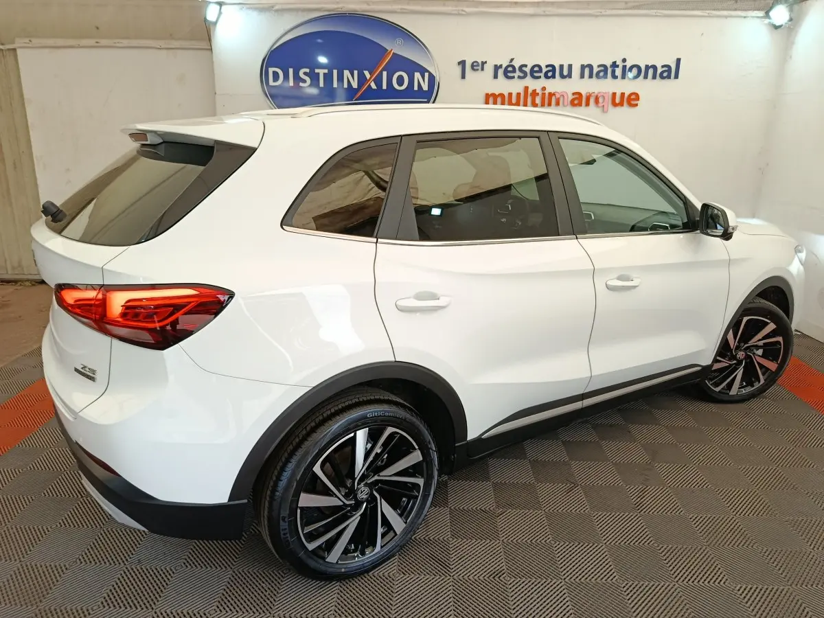 Vue 3/4 arrière droite du MG ZS blanc 2026 avec jantes noires et feux arrière LED allumés dans un showroom.
