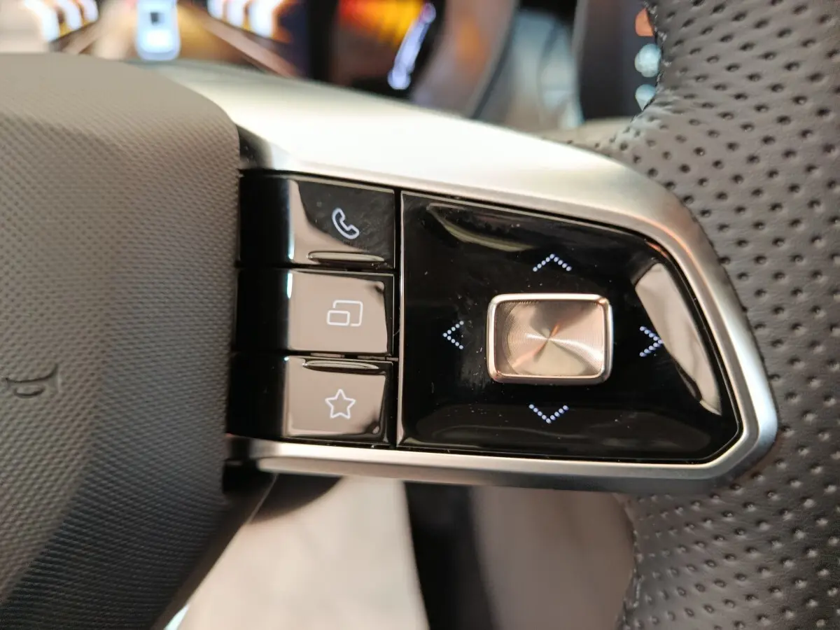 Gros plan sur les commandes gauche du volant perforé noir du MG ZS blanc, avec boutons multifonctions et finition argentée.
