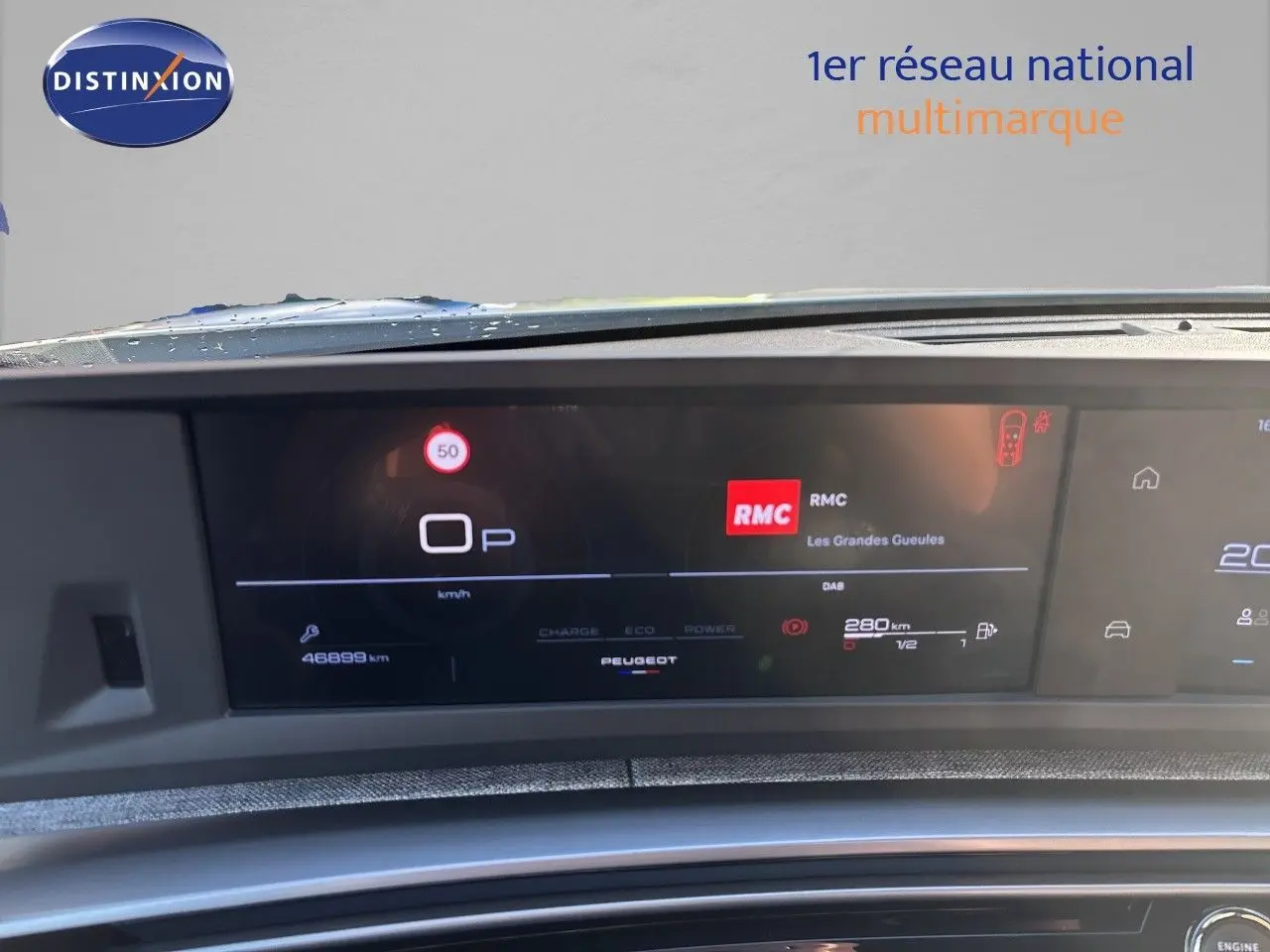 Tableau de bord numérique du Peugeot 5008 blanc, affichant la vitesse, la radio RMC et l'autonomie.