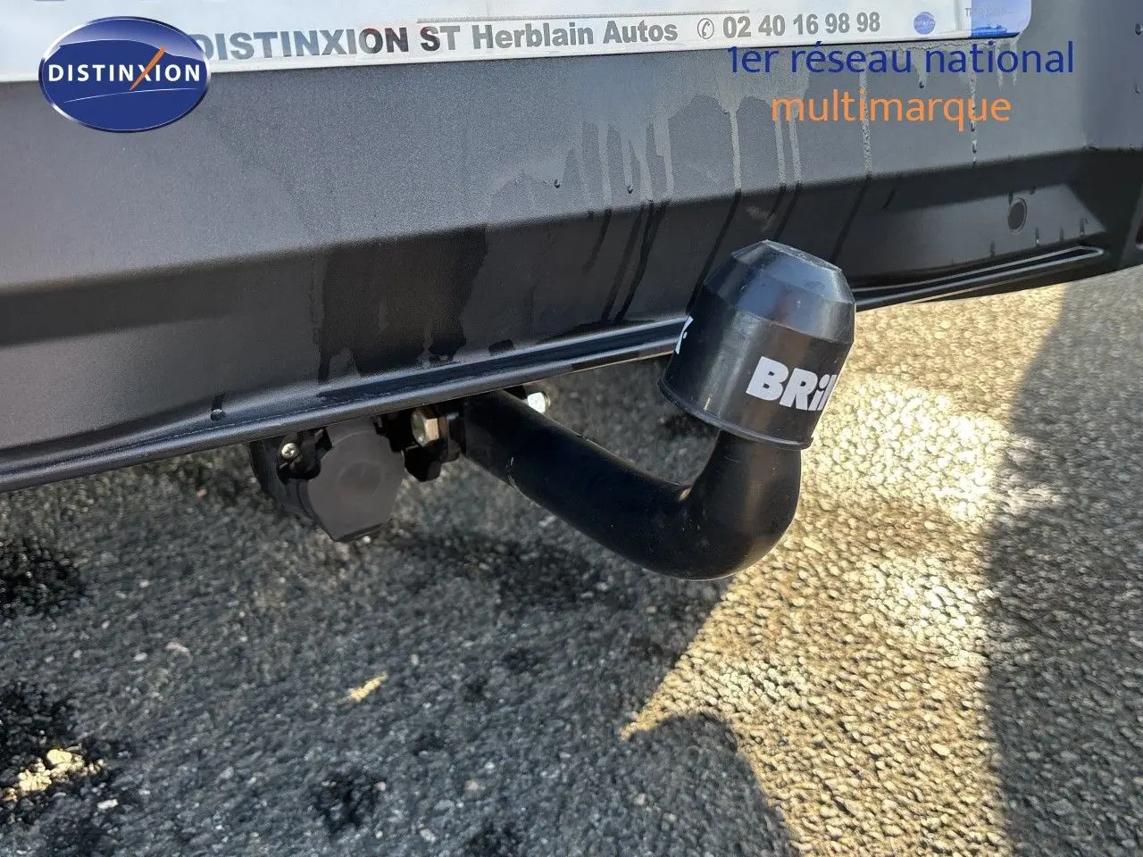 Gros plan sur l'attelage noir fixé à l'arrière d'un Peugeot 5008 blanc Okenite, avec fond de sol goudronné.
