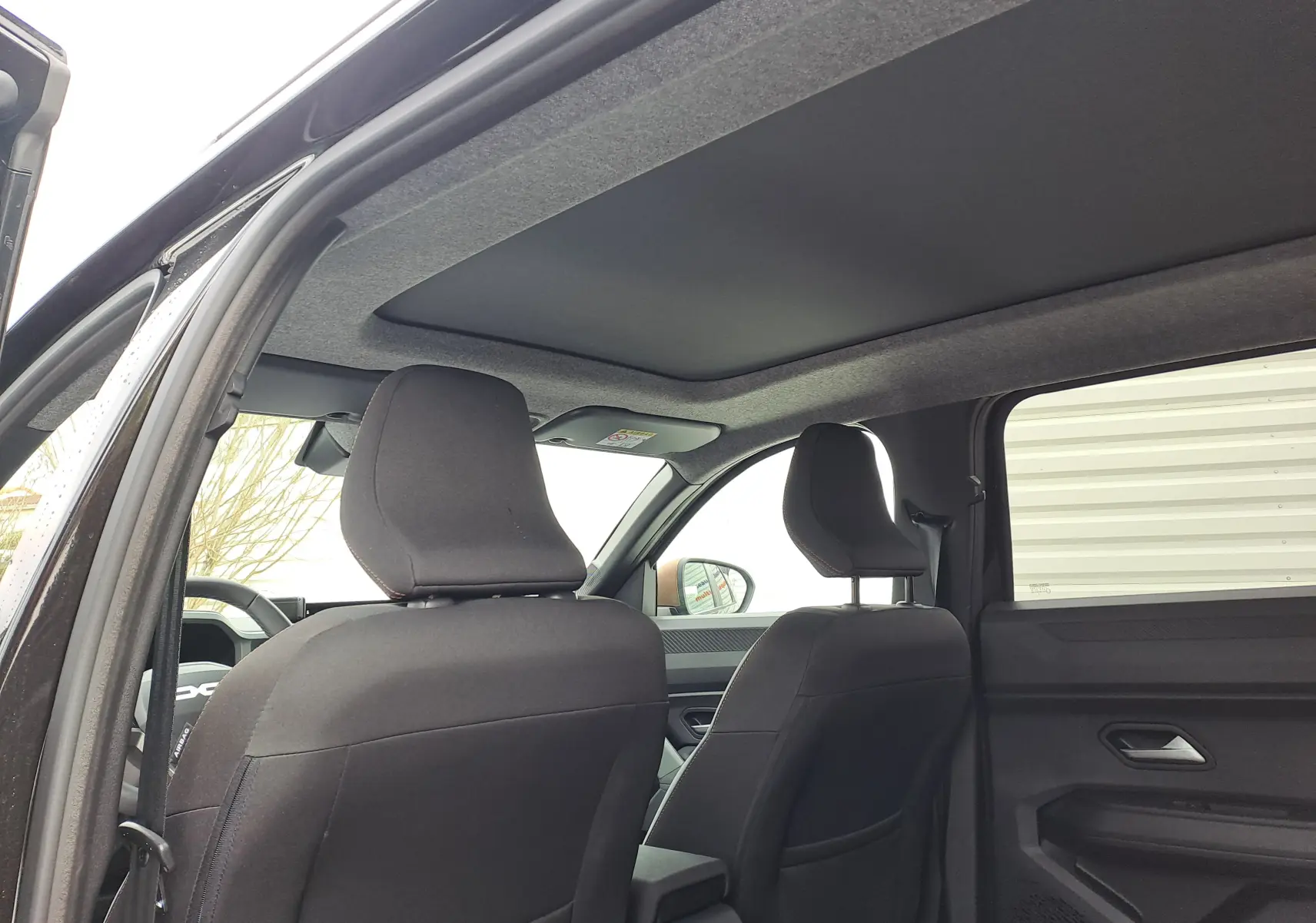 Vue intérieure arrière montrant les sièges avant en tissu gris et le toit noir nacré du Dacia Bigster 2025.