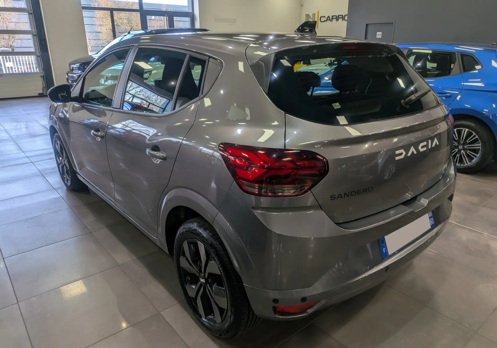 Vue 3/4 arrière droite d'une Dacia Sandero TCe 90 gris schiste en showroom, avec jantes noires distinctives.