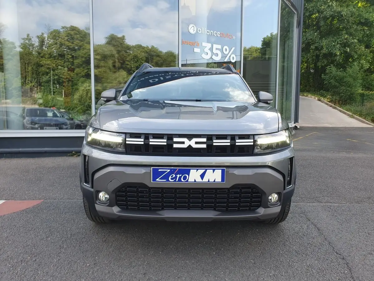 Vue avant d'un Dacia Duster 2026 gris schiste avec calandre noire et phares allumés, devant une vitrine de concession.
