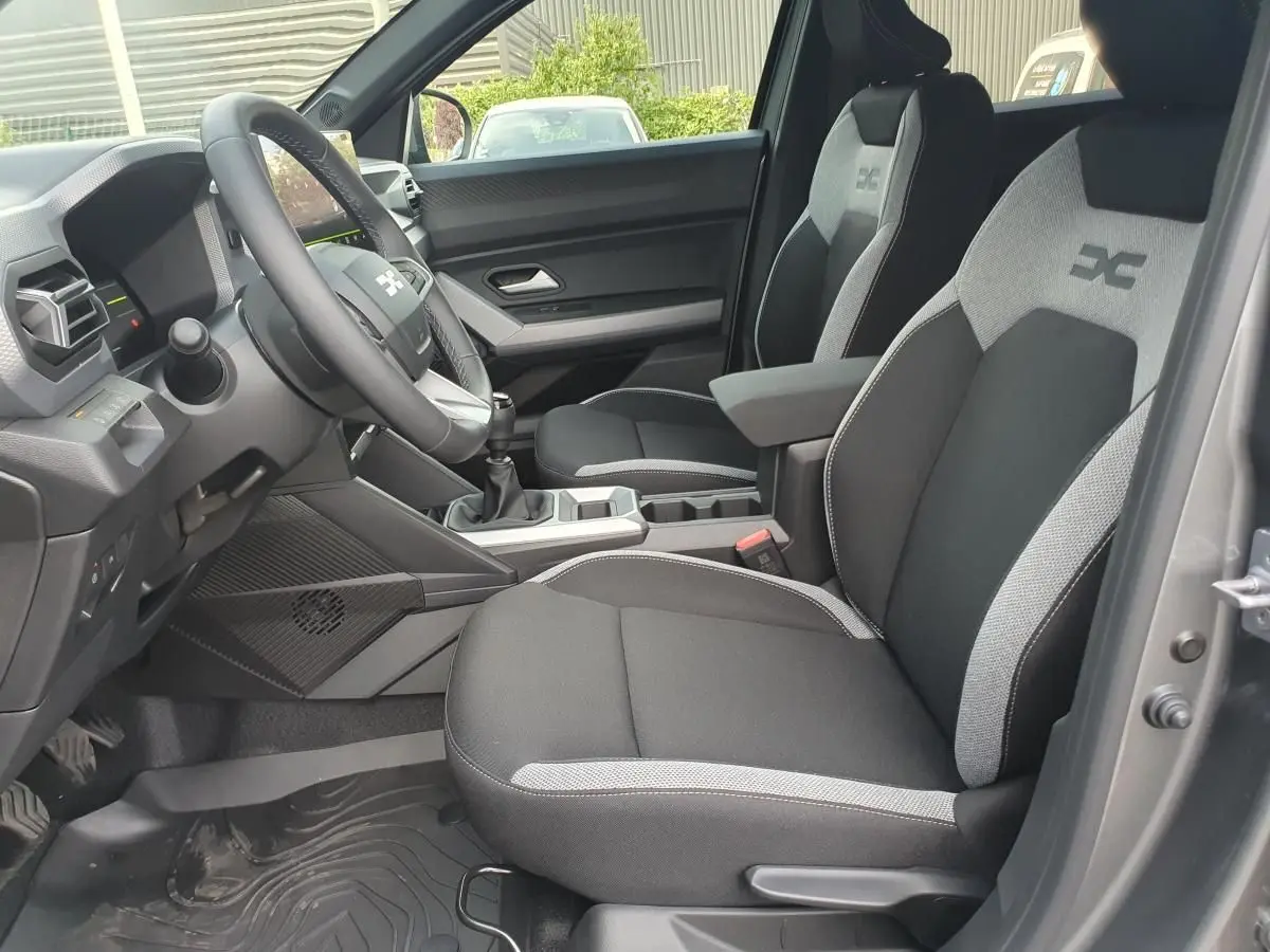 Vue intérieure côté conducteur du Dacia Duster 2026, sièges tissu noir et gris avec logo distinctif brodé.