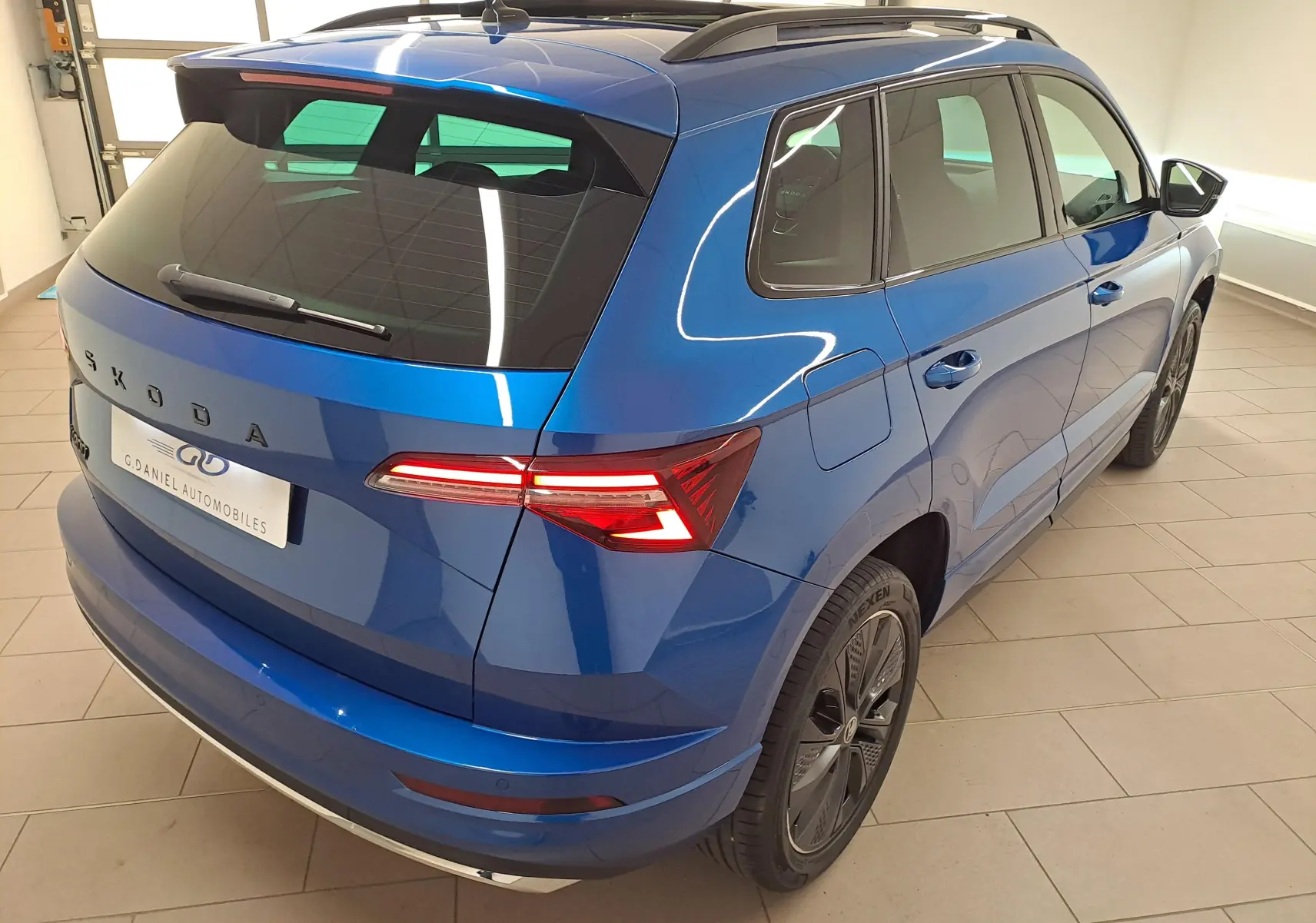 Vue 3/4 arrière droite d'un Skoda Karoq 2025 Bleu RS avec toit panoramique et feux LED allumés dans un showroom.