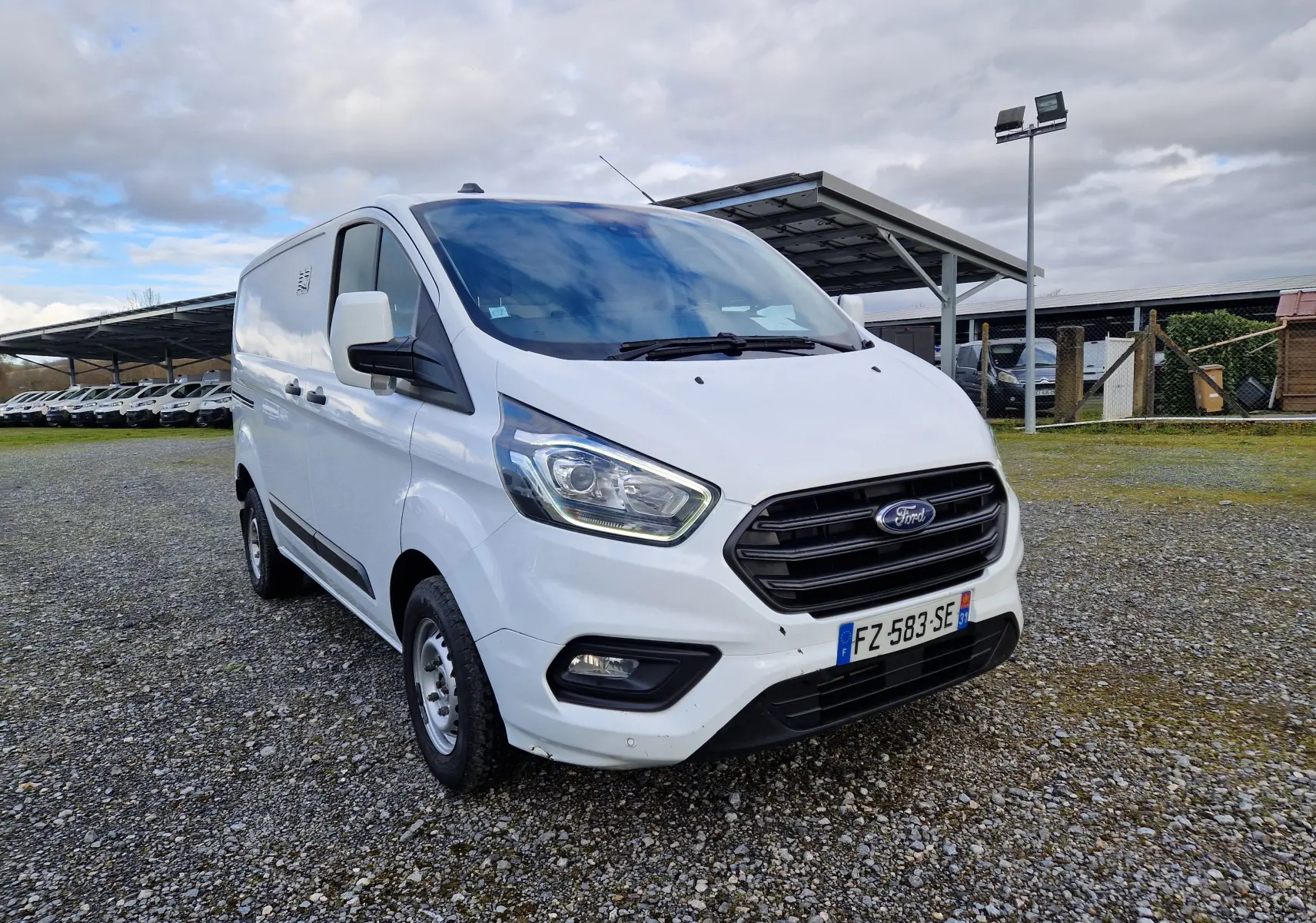Ford Transit Custom blanc en vue 3/4 avant droit sur un sol gravillonné avec un ciel nuageux en arrière-plan.