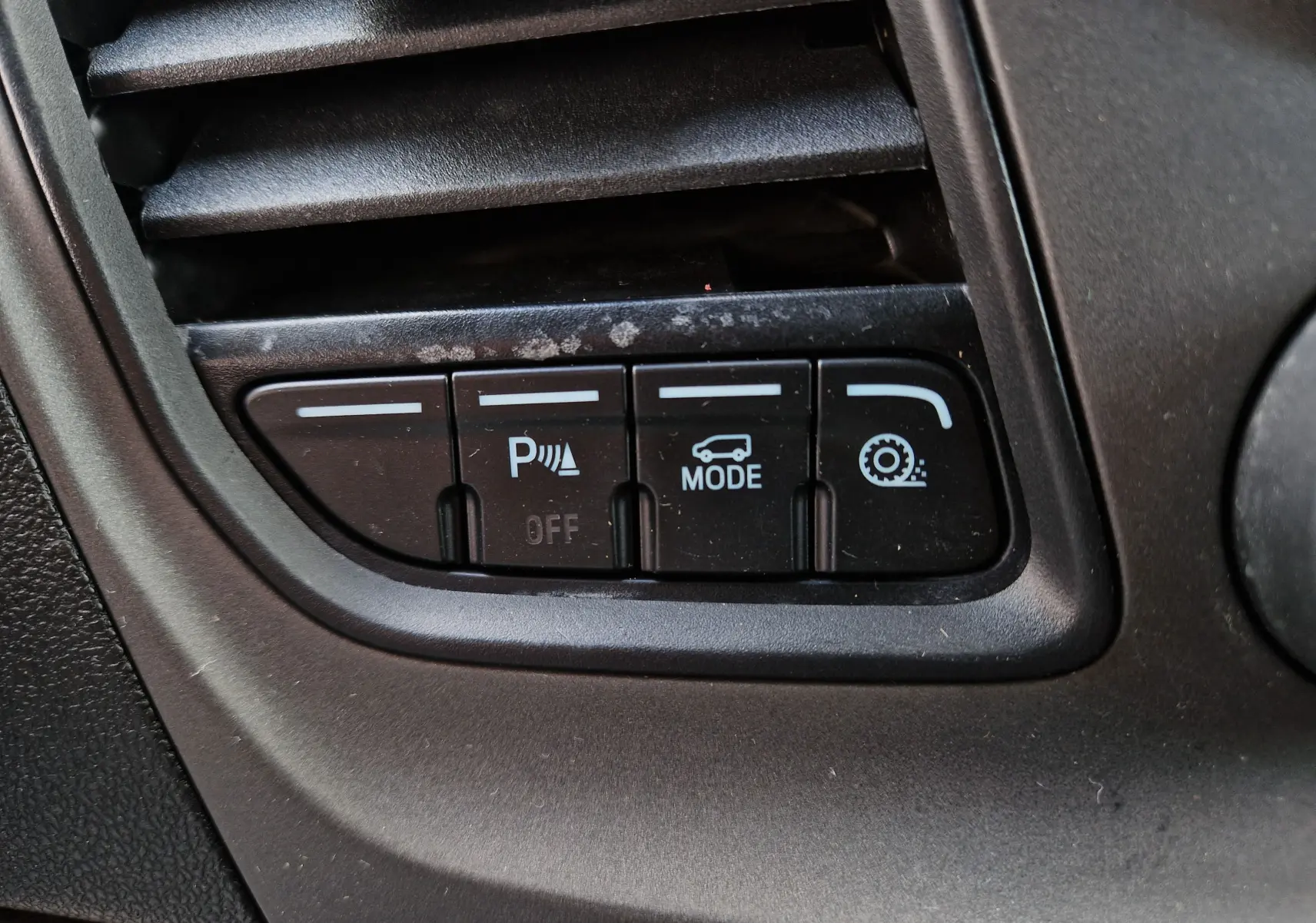 Gros plan sur les boutons de commande noirs du tableau de bord du Ford Transit Custom blanc, avec icônes de parking, mode et régulateur.