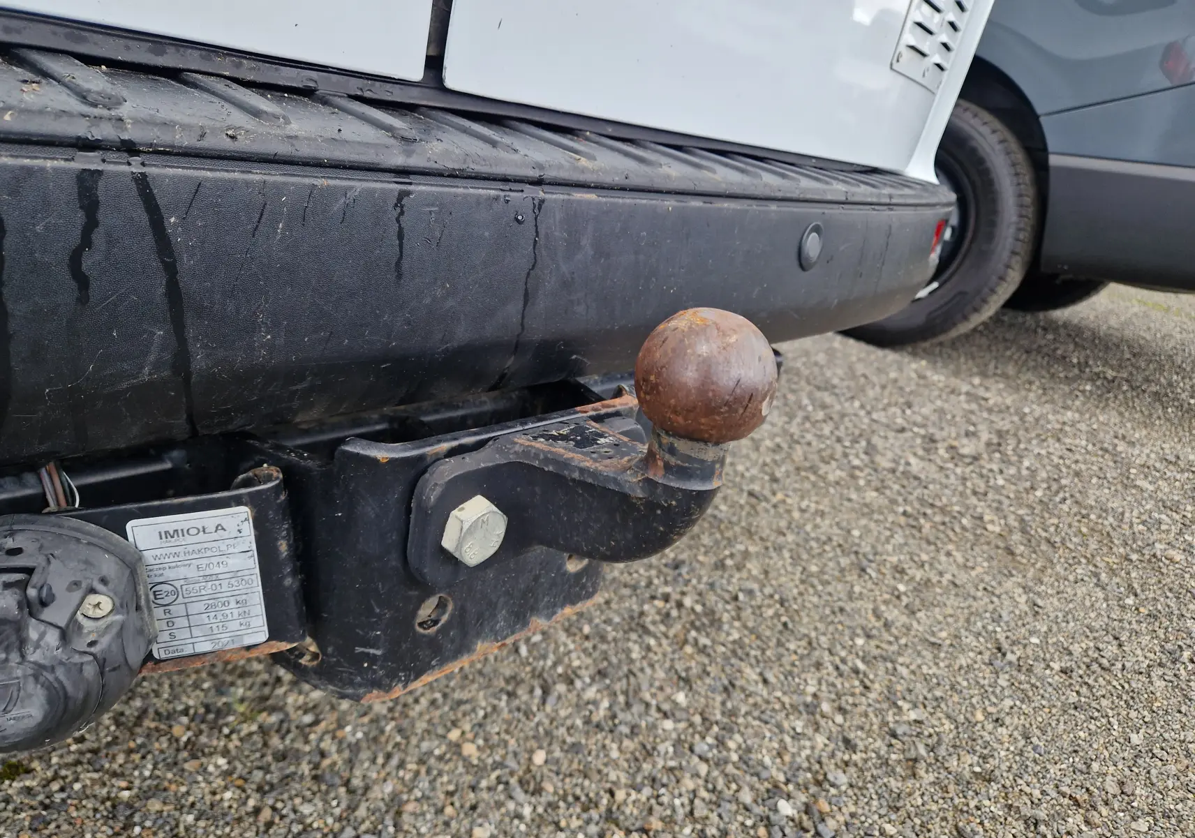 Gros plan sur l'attelage rouillé à boule fixé au pare-chocs arrière noir du Ford Transit Custom blanc, vue côté gauche.