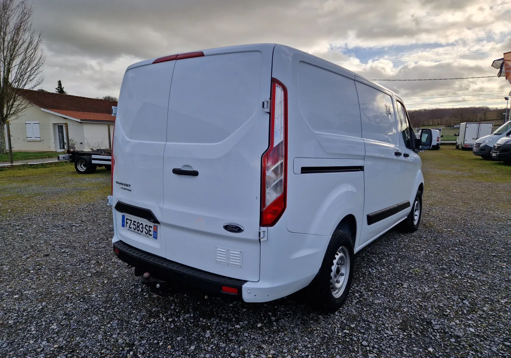Vue 3/4 arrière droite d'un fourgon Ford Transit Custom blanc, avec portes arrière fermées et plaque d'immatriculation visible.