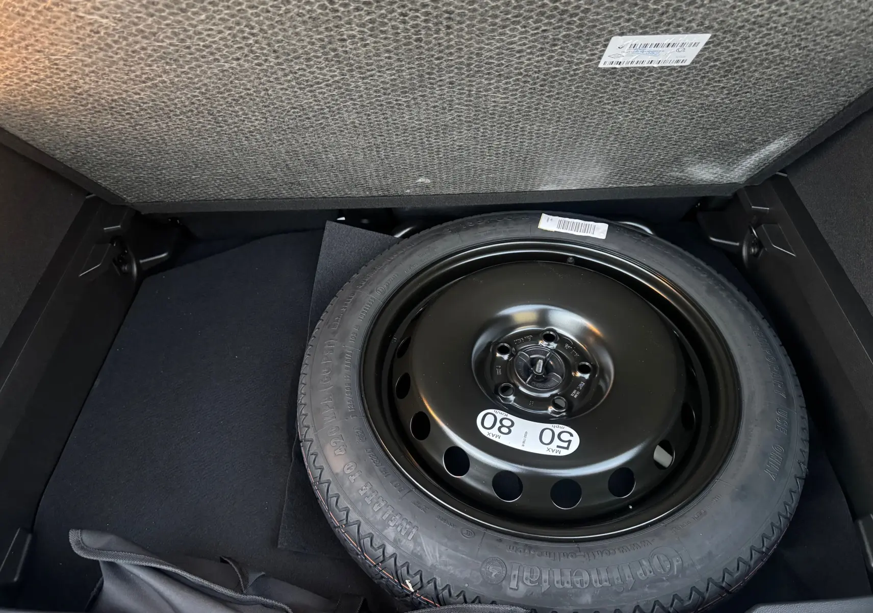 Roue de secours noire dans le coffre d’un Renault Captur blanc nacré toit noir, vue en plongée sur le pneu.