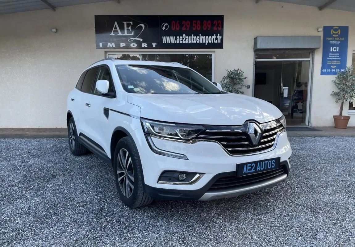 Renault Koleos blanc vu en 3/4 avant droit, avec calandre chromée et jantes alliage, stationné devant un bâtiment.