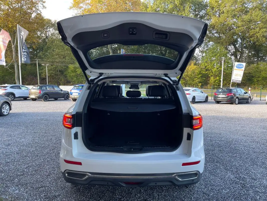 Vue arrière d'un Renault Koleos blanc 2019 avec coffre ouvert, montrant l'espace de chargement et les feux arrière allumés.