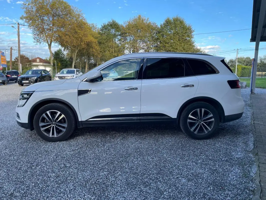 Renault Koleos blanc vu de profil côté gauche, SUV avec jantes alliage et vitres teintées, stationné sur gravier.