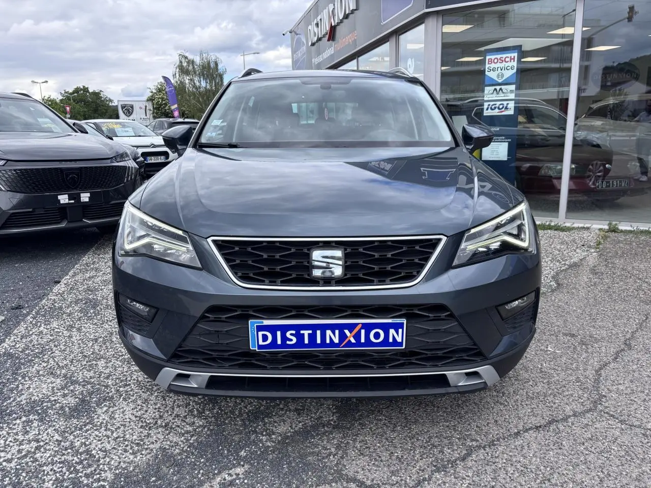 Vue de face d'un SEAT Ateca gris foncé avec calandre noire chromée et phares LED, stationné devant un garage.