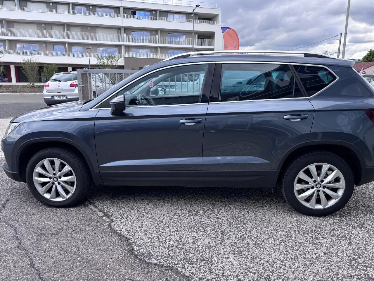 Vue latérale droite d'un SEAT Ateca gris foncé 2019 avec jantes alliage 17'' et barres de toit chromées.