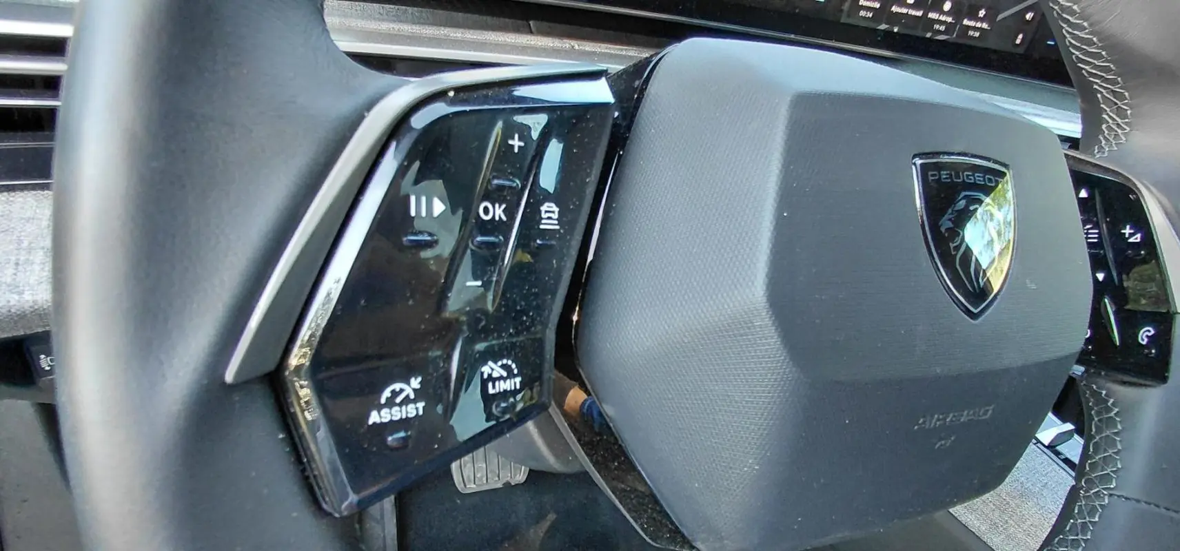 Gros plan sur le volant gris du Peugeot 5008 Hybrid 2025, avec commandes multifonctions et logo central visible.