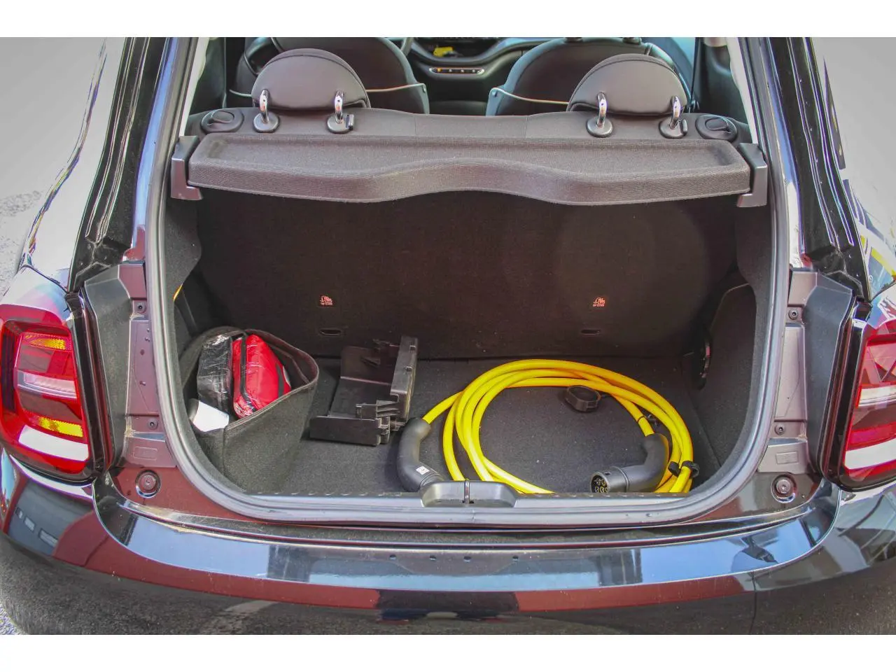 Coffre ouvert vu de l'arrière d'une Fiat 500 électrique noire avec câble de recharge jaune et accessoires rangés.