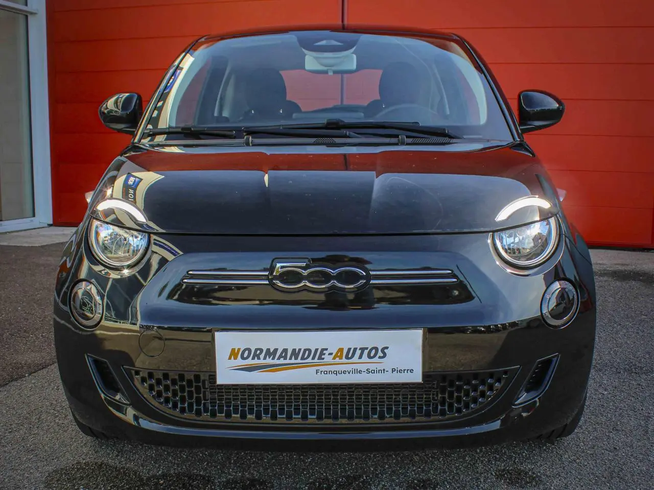 Vue frontale d'une Fiat 500 électrique noire avec logo 500 chromé sur calandre, devant un mur rouge.