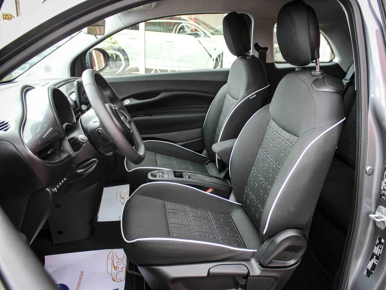 Vue intérieure côté conducteur de la Fiat 500 électrique 2023, sièges tissu noir avec surpiqûres blanches et tableau de bord noir.