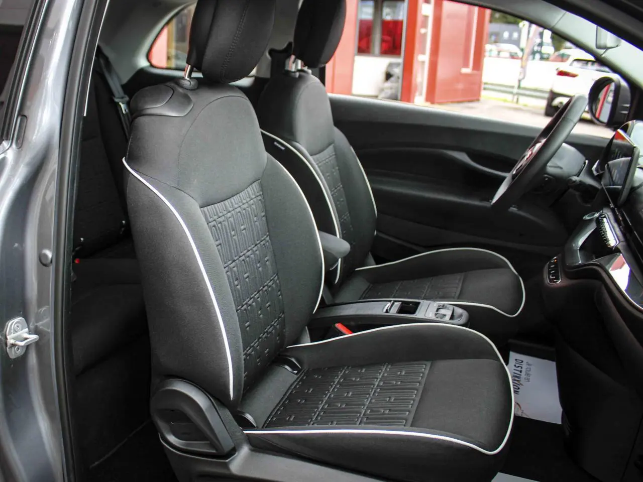 Vue intérieure côté conducteur de la Fiat 500 électrique 2023, sièges tissu noir avec surpiqûres blanches et console centrale.