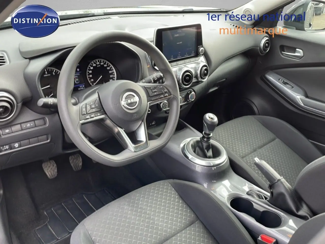 Vue intérieure de face côté conducteur du Nissan Juke 2023, volant multifonction et levier de vitesse manuel, finition tissu gris-noir.