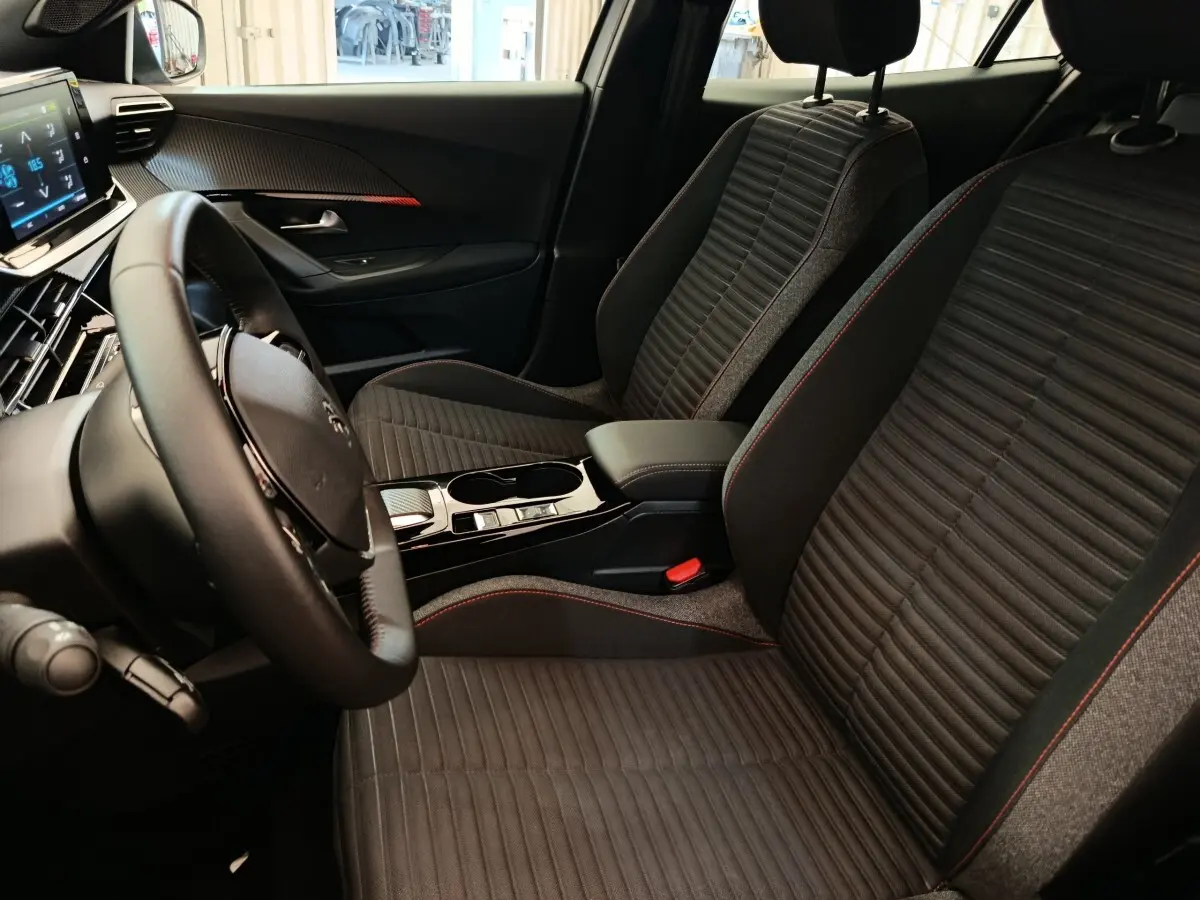 Vue intérieure côté conducteur du Peugeot E-2008 électrique, sièges noirs avec surpiqûres rouges et volant cuir multifonctions.
