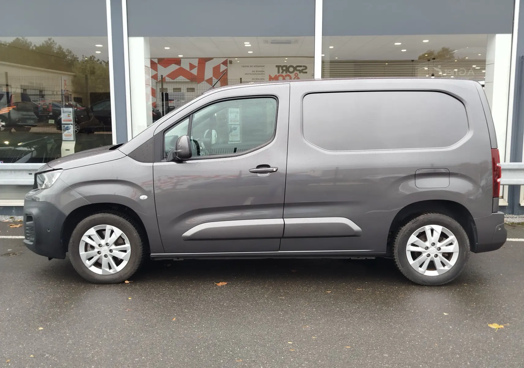 Vue de profil côté gauche d'un Peugeot Partner gris foncé stationné devant une vitrine de concession.