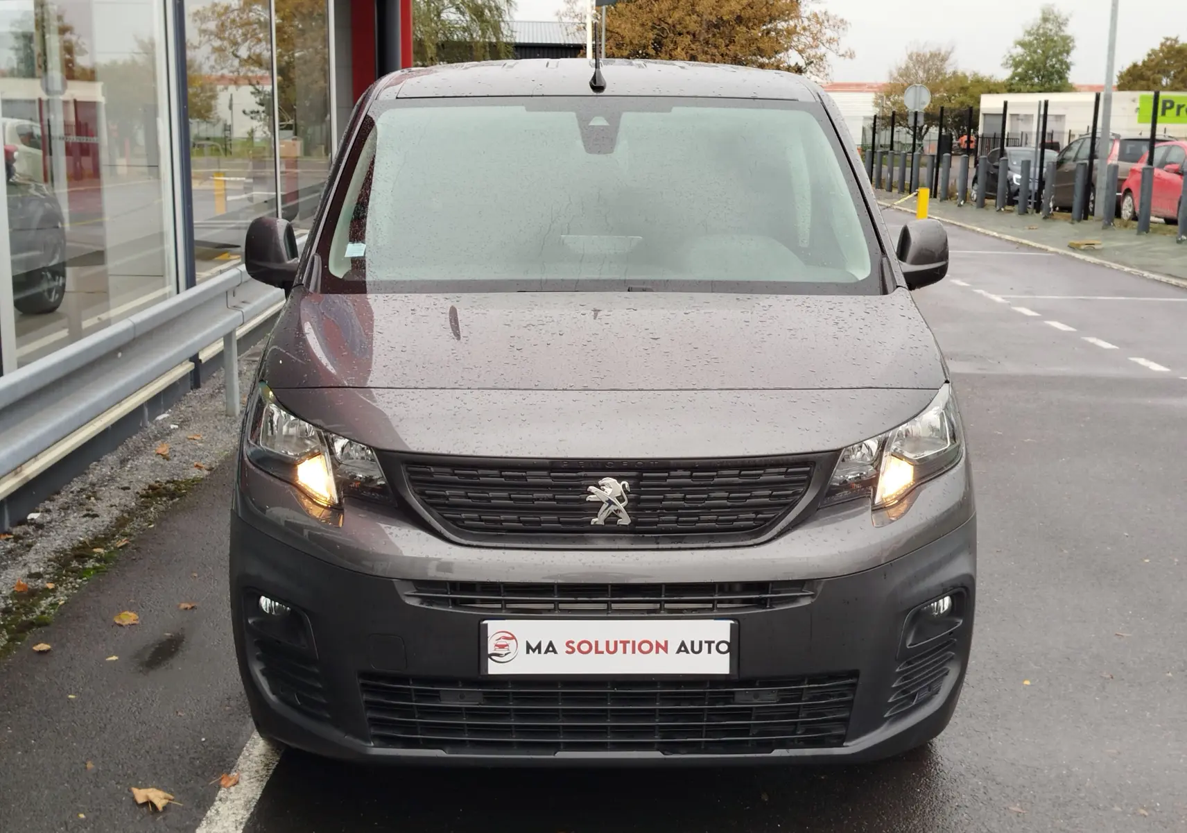 Vue de face d’un Peugeot Partner gris foncé 2020 avec feux avant allumés sur un parking humide.