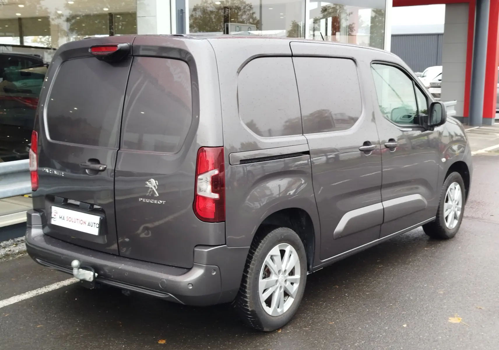 Vue 3/4 arrière droite d'un Peugeot Partner gris foncé, utilitaire compact avec portes latérales pleines et attelage visible.