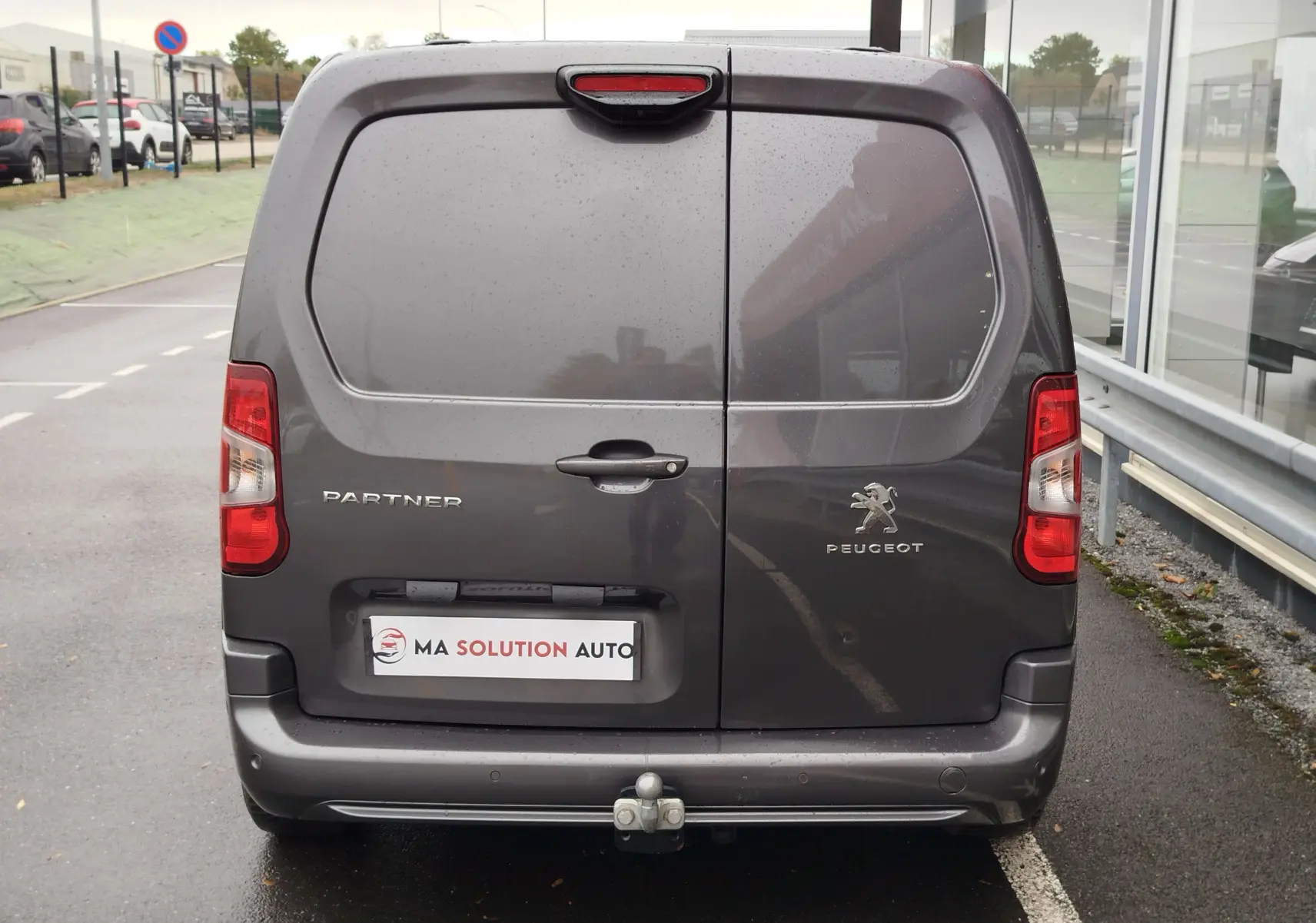 Vue arrière d’un Peugeot Partner gris foncé 2020 avec portes battantes et attelage visible sur un parking humide.