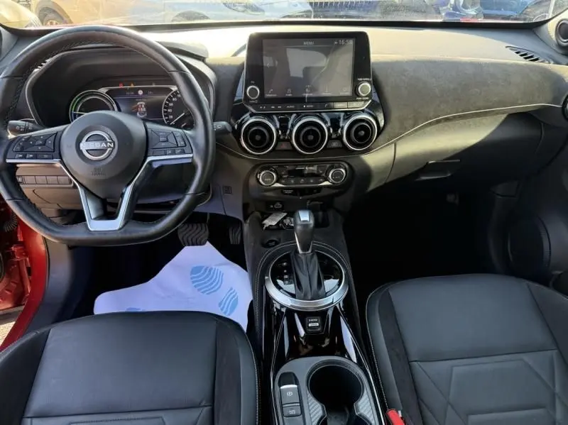 Vue intérieure avant du Nissan Juke 1.6 Hybrid 2024 rouge Fuji, tableau de bord avec écran tactile et volant multifonction noir.