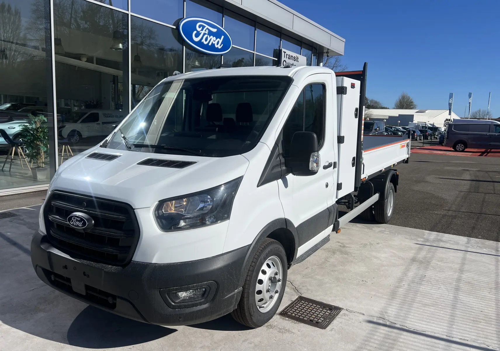 Vue 3/4 avant droit d’un Ford Transit Chassis Cabine blanc avec benne et coffre, roues jumelées, stationné devant un showroom Ford.