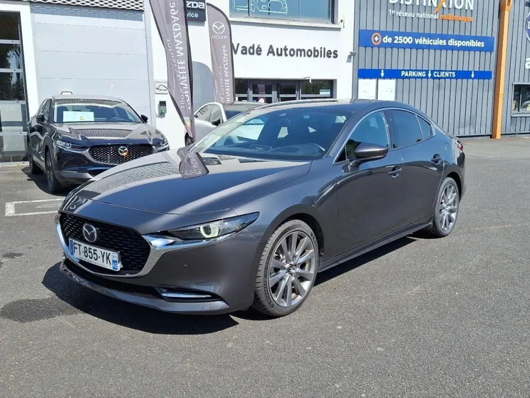 Vue 3/4 avant d'une Mazda3 gris métallisé garée devant un concessionnaire avec jantes aluminium et phares allumés.