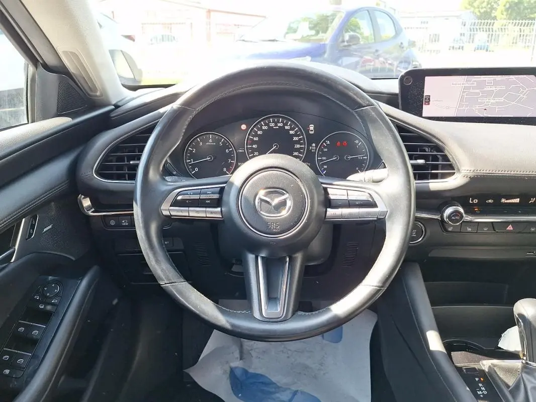 Vue intérieure centrée sur le volant cuir multifonctions d'une Mazda3 gris, avec tableau de bord et écran GPS visibles.