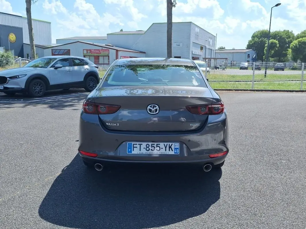 Vue arrière d'une Mazda3 gris métallisé stationnée, avec feux arrière allongés et double sortie d'échappement visible.