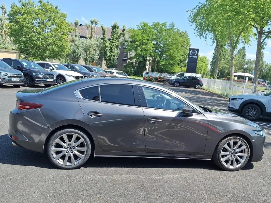 Vue de profil côté droit d'une Mazda3 gris métallisé stationnée sur un parking extérieur, avec jantes aluminium visibles.