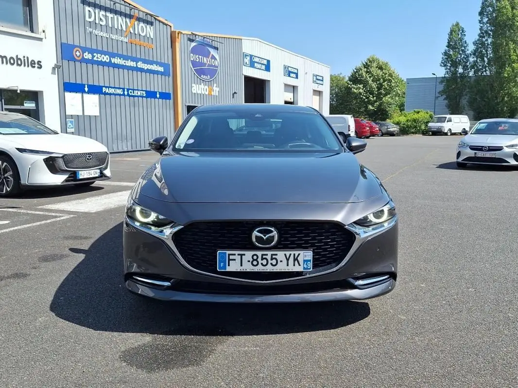 Vue avant d'une Mazda3 gris métallisé avec calandre noire et phares LED allumés, stationnée devant un bâtiment commercial.