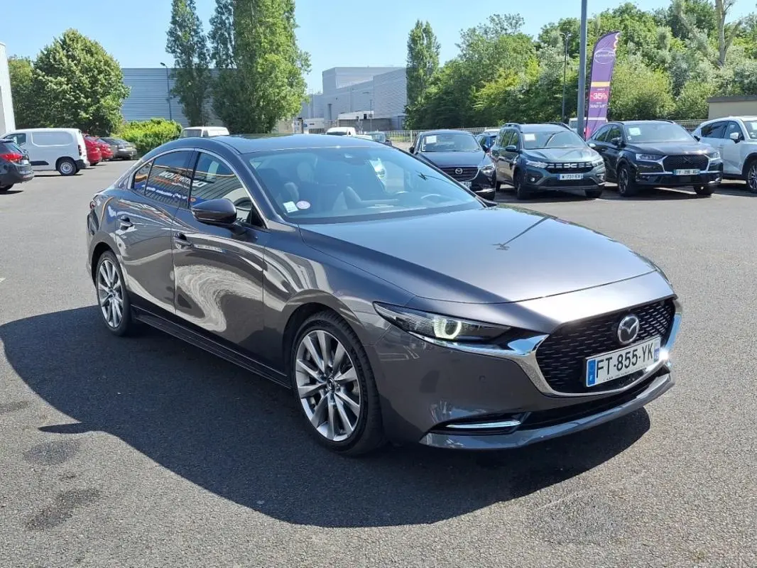 Vue 3/4 avant d'une Mazda3 gris métallisé 4 portes, avec phares allumés et jantes alliage visibles.