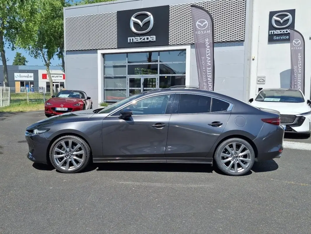 Vue latérale droite d'une Mazda3 gris métallisé garée devant une concession Mazda avec jantes alu et vitres teintées.