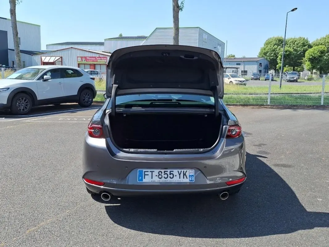 Vue arrière d'une Mazda3 gris métallisé avec coffre ouvert sur un parking extérieur ensoleillé.