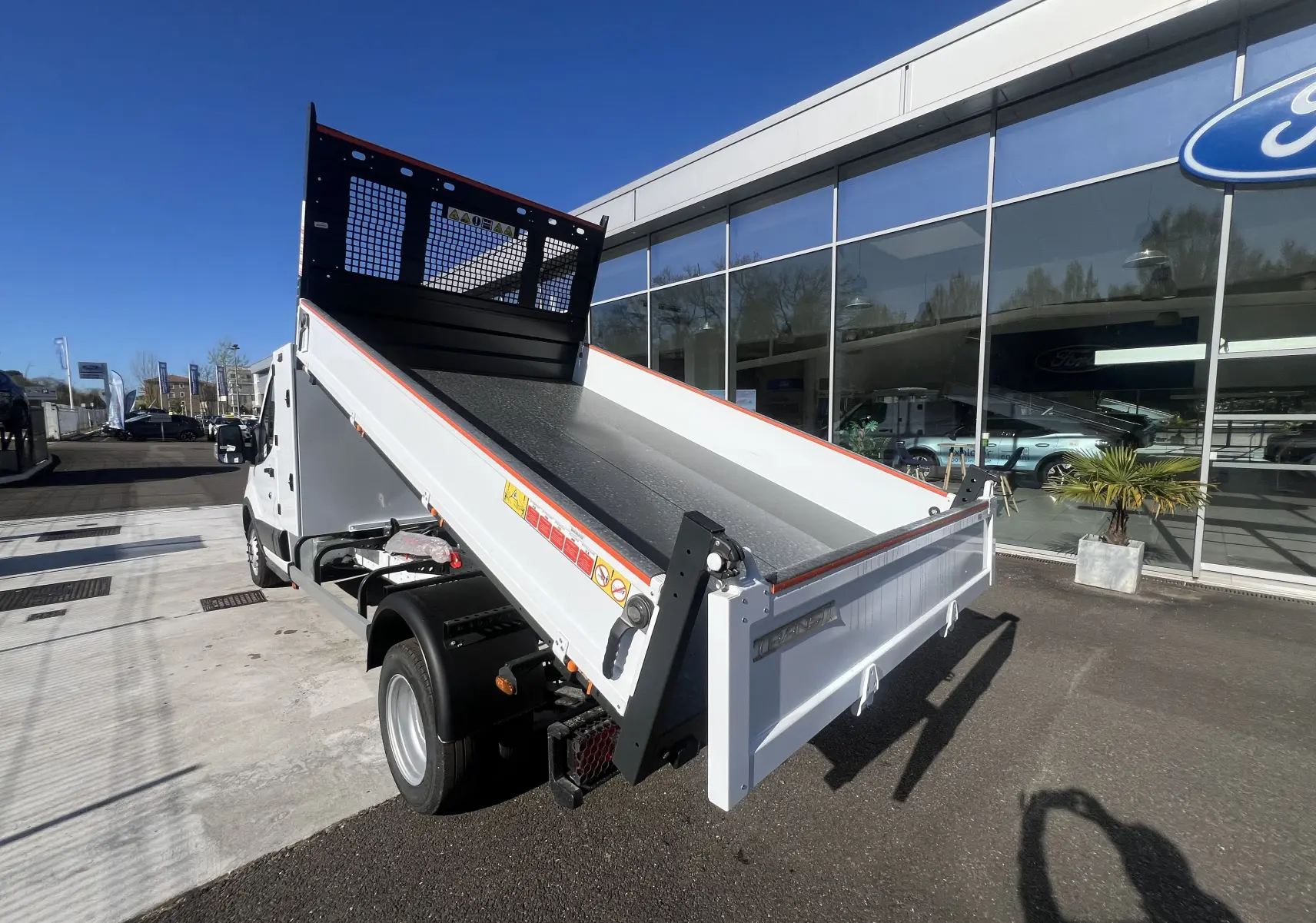 Vue 3/4 arrière droite du Ford Transit Chassis Cabine blanc avec benne relevée et coffre, roues jumelées visibles.
