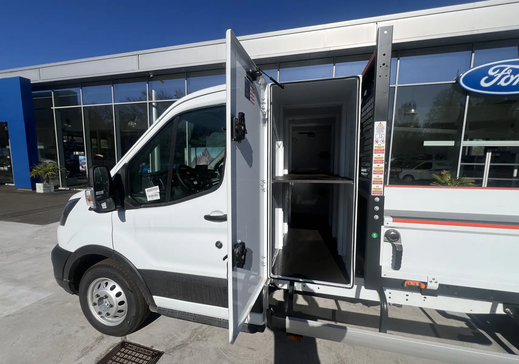 Vue de profil côté gauche d’un Ford Transit blanc avec porte latérale ouverte révélant un coffre de rangement intégré à la benne.
